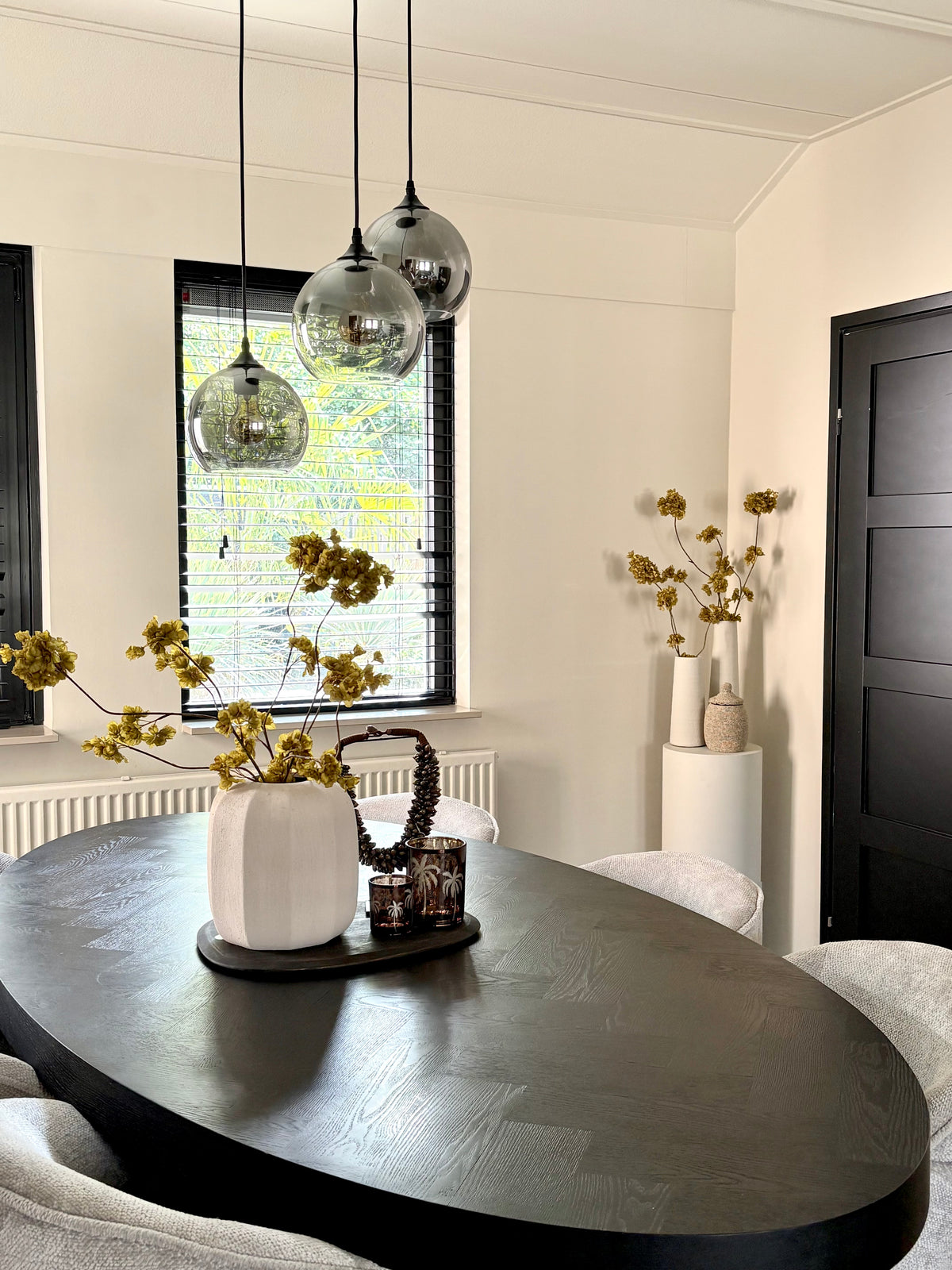 Een woonkamer met een setje op de eettafel met een crème vaas, olijfgroene bloesemtakken en bruine woonaccessoires